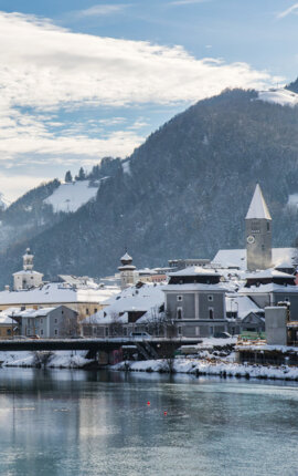 Winterliche Ansicht von Hallein: verschneite Dächer, Kirchturm und Brücke an der Salzach vor schneebedeckten Bergen.
