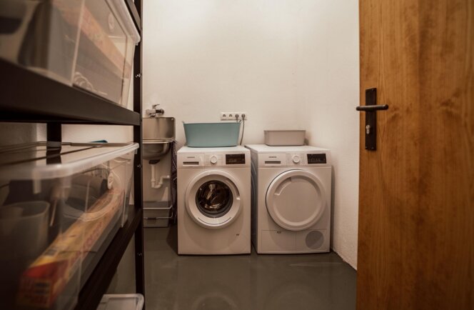 Guest laundry room with a washing machine and dryer, storage shelves with plastic bins on the left, and a small sink in the corner.