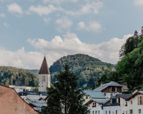 Blick auf die Stadt Hallein mit Kirchturm vor bewaldeten Hügeln und blauem Himmel.