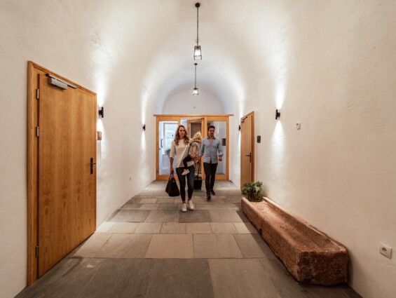 Personen betreten einen Flur mit gewölbter Decke und Holztüren in einem stilvollen Gebäude.