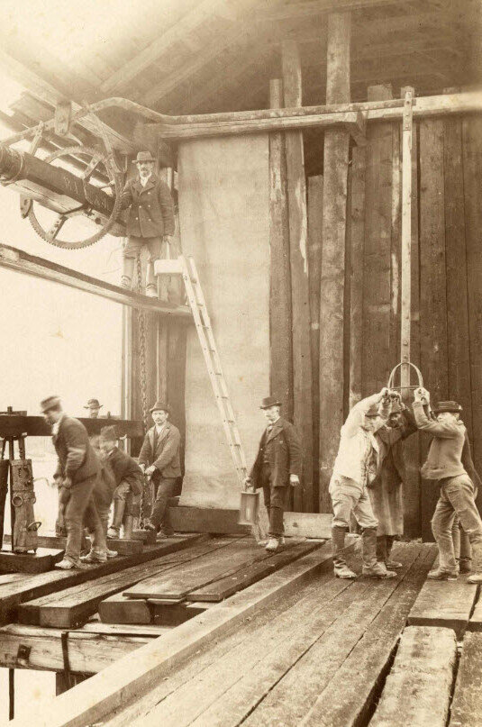 Arbeiter auf einer Baustelle mit Maschinen und Holzstrukturen, historische Aufnahme.