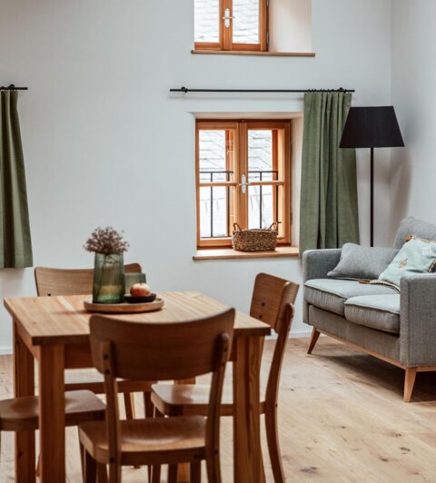A cozy living room with a wooden dining table, green curtains, a gray sofa, and wooden flooring, creating a warm and inviting atmosphere.