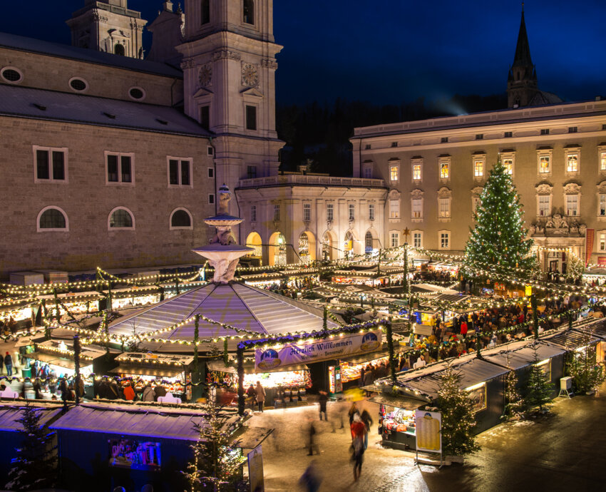 Stimmungsvoller Christkindlmarkt am Salzburger Domplatz bei Nacht, mit Lichterketten, Ständen und großem Baum.