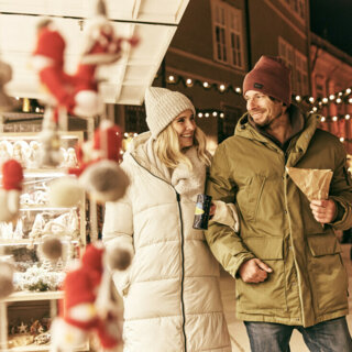 Ein Paar spaziert über einen stimmungsvollen Weihnachtsmarkt in Hallein und genießt Punsch und Maroni unter Lichterketten.