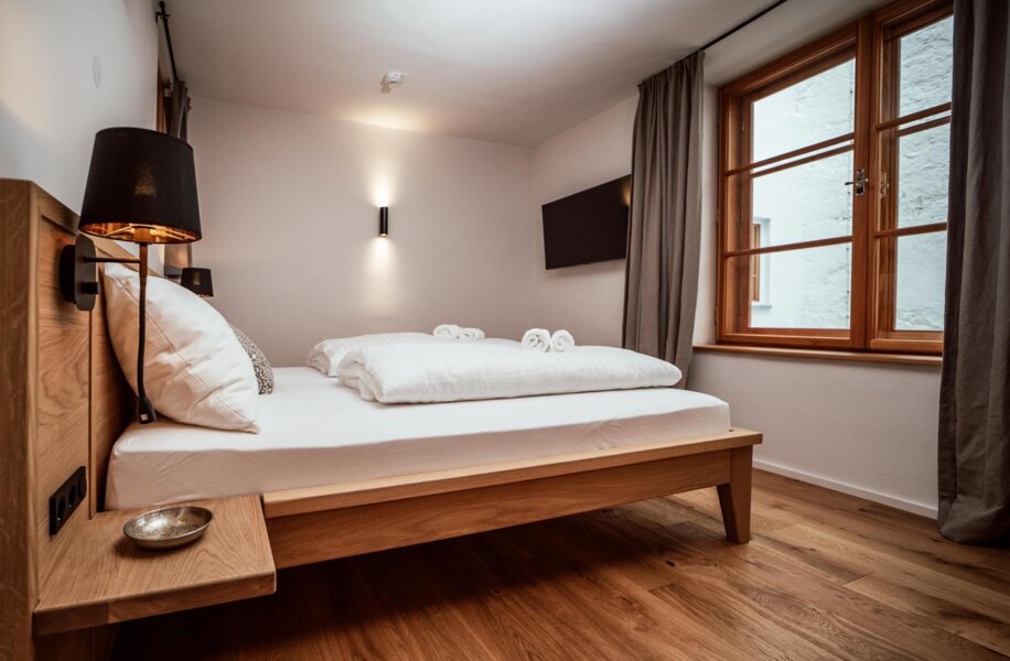 Cozy bedroom with a wooden bed, white bedding, and a window with curtains. Modern wall lights and a small bedside shelf add to the inviting atmosphere.