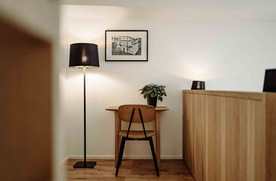 Ein Arbeitsbereich mit Holzstuhl, Stehlampe und Kunstwerk an der Wand. Ein Topf mit Blumen steht auf dem Tisch.