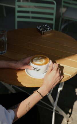 Eine Person hält eine Tasse Kaffee mit Latte Art auf einem Holztisch im Freien.