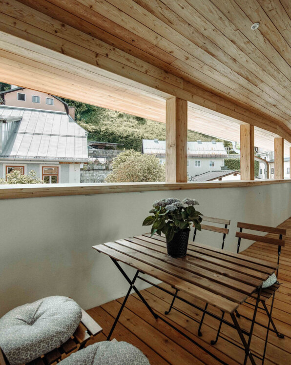 Wooden balcony with a table and chairs, overlooking neighboring houses and greenery. Cozy and inviting outdoor space.