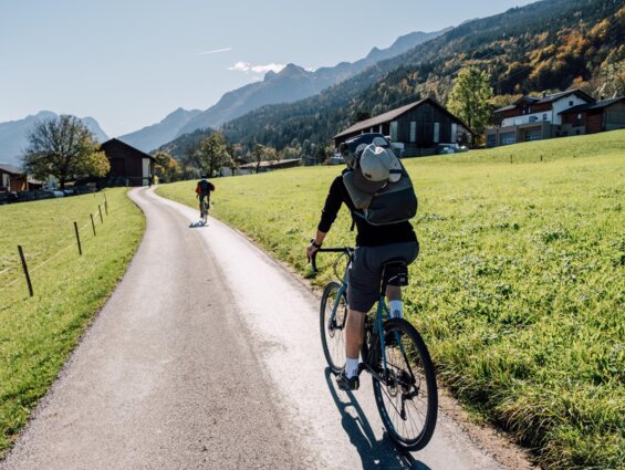 Zwei Radfahrer auf einem ländlichen Weg in einer malerischen Berglandschaft, mit grünen Wiesen und traditionellen Häusern.