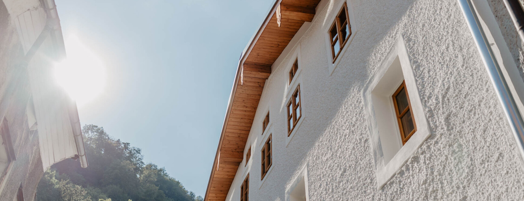 Weiße Außenfassade von the salt in Hallein mit Holzfenstern unter blauem Himmel, Sonne scheint von links, umgeben von grünen Bäumen.