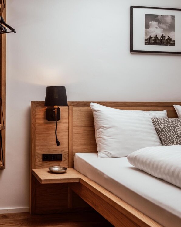 Cozy bedroom with wooden bed frame, white bedding, and a black lamp. A framed photo hangs on the wall above a wooden nightstand.
