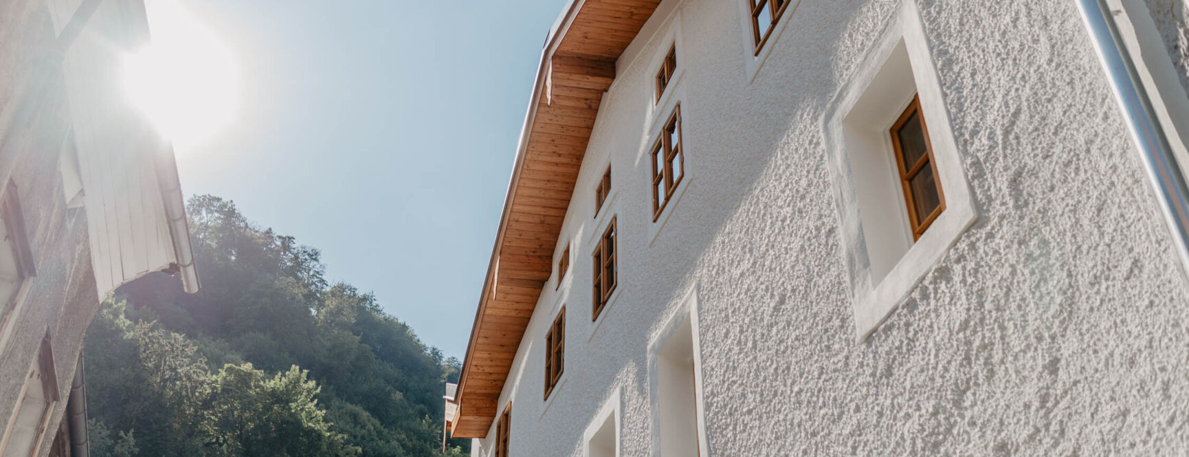 Sunlit building with textured white walls and wooden windows, nestled against a backdrop of green trees and clear blue sky.