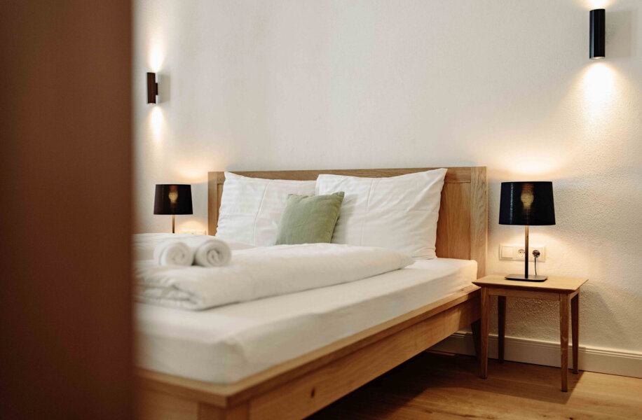 Ein gemütliches Schlafzimmer mit Holzbett, weißen Bettwäsche, grünen Kissen und Nachttischen mit schwarzen Lampen in einem Hotelzimmer.