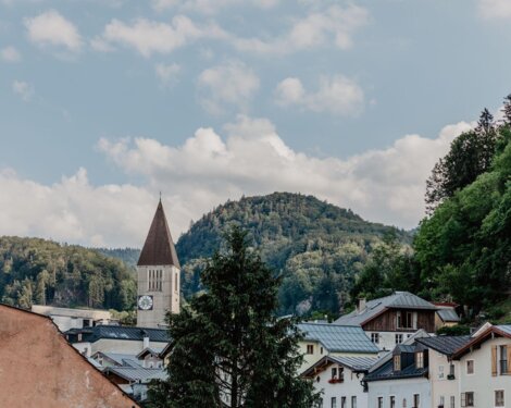 Blick auf die Stadt Hallein mit Kirchturm vor bewaldeten Hügeln und blauem Himmel.