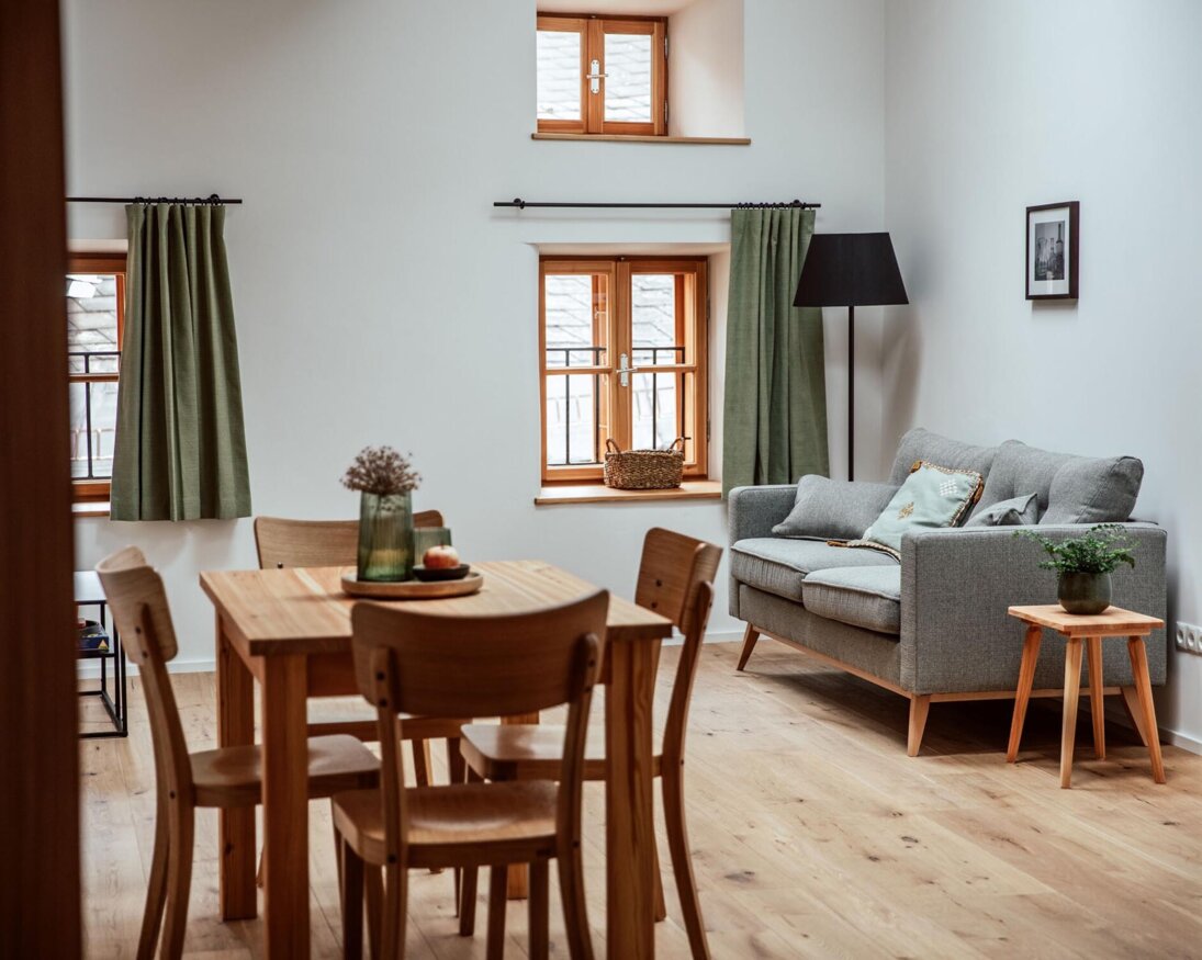 Gemütlicher Wohnbereich in einem Apartment von the salt Hallein in Salzburg mit Holztisch, grauem Sofa und grünen Vorhängen.