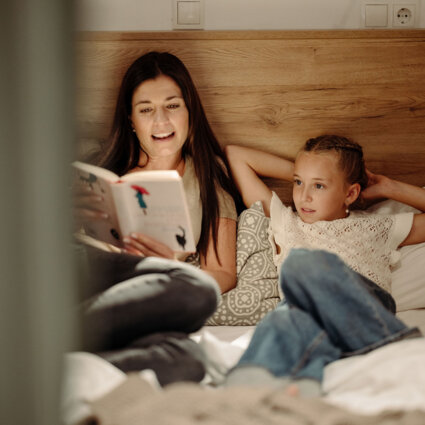 Eine erwachsene Person liest einem Kind im Bett vor. Warmes Leselicht, gemütliches Zimmer im the salt Hallein.