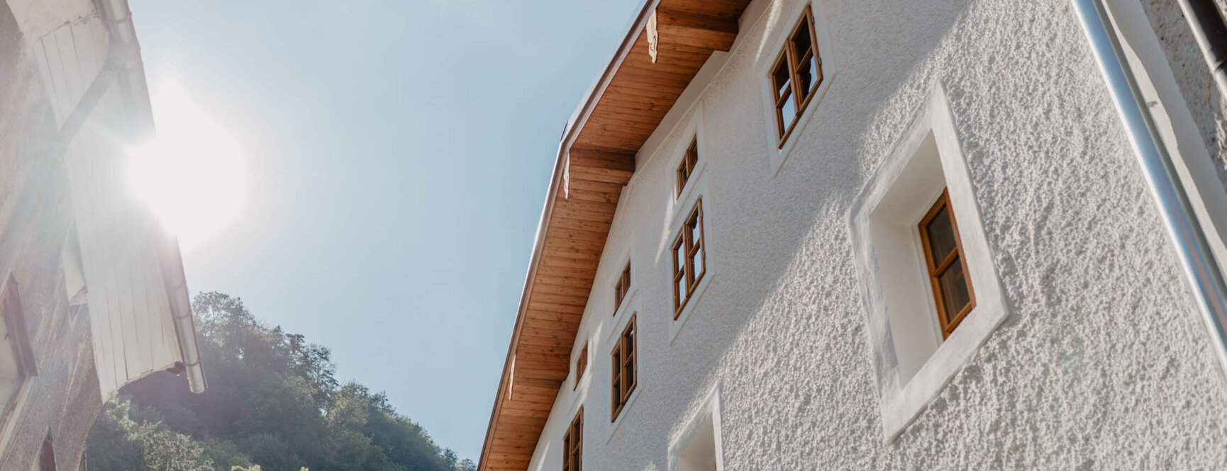 Weiße Außenfassade von the salt in Hallein mit Holzfenstern unter blauem Himmel, Sonne scheint von links, umgeben von grünen Bäumen.
