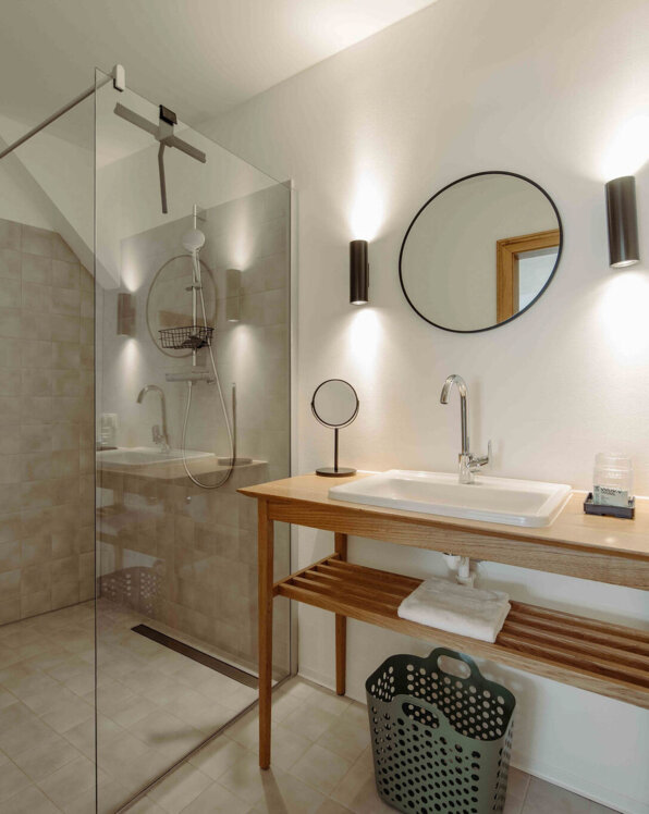 Modern bathroom with a glass shower, wooden vanity, round mirror, and soft lighting.