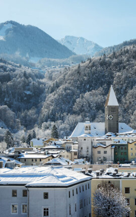 Snow-covered Hallein with colorful buildings and a church tower, nestled in a mountainous landscape under a clear blue sky.