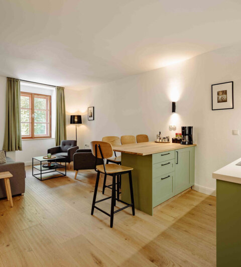 Modern living area with a cozy sitting space, wooden flooring, and a small kitchen with light green cabinets in a well-lit room.