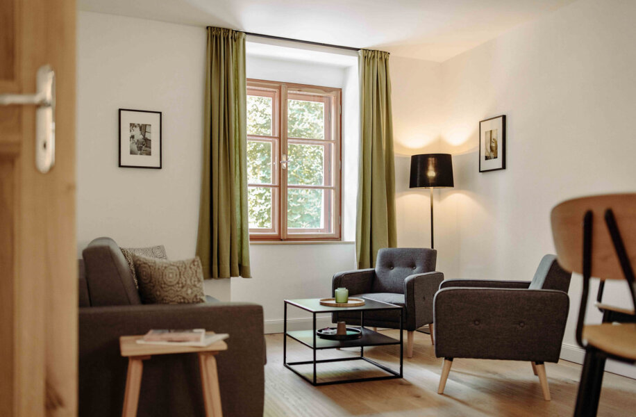 Gemütliches Wohnzimmer in einer stilvollen Ferienwohnung in Salzburg mit grauen Sesseln, Holztisch und grünen Vorhängen.