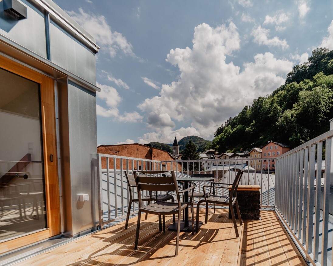 Eine Terrasse von einem der Rooftop-Apartments mit Tisch und Stühlen, umgeben von grünen Hügeln und bunten Gebäuden unter einem blauen Himmel mit Wolken.