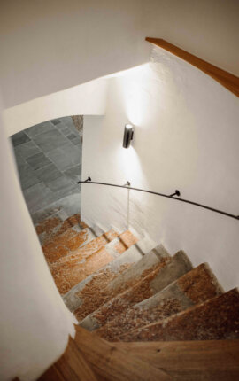 Eine geschwungene Treppe mit rustikalen Steinfliesen und Wandbeleuchtung führt nach unten.