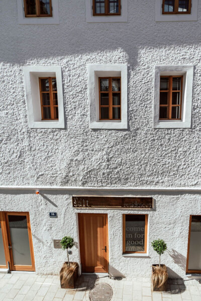 Facade of a white building with wooden windows and doors, featuring a sign reading "Salt" above the entrance. Potted plants flank the doorway.