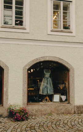Schaufenster eines Bekleidungsgeschäfts mit einem Trachtenkleid auf einer Puppe, darunter ein Blumenarrangement.
