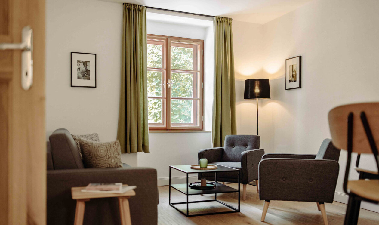 Gemütliches Wohnzimmer in einer stilvollen Ferienwohnung in Salzburg mit grauen Sesseln, Holztisch und grünen Vorhängen.