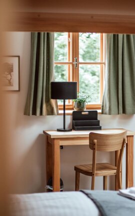 Gemütlicher Schreibtischbereich im Hotelzimmer mit Holzstuhl, Tischlampe und Fenster mit grünen Vorhängen.