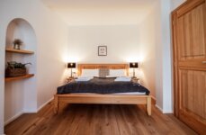 Gemütliches Schlafzimmer mit Holzbett, weißen Wänden und Holzboden. Zwei Lampen auf Nachttischen. Regal mit Pflanzen links.