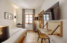 Modernes Hotelzimmer mit Einzelbett, Schreibtisch, Flachbildfernseher und großem Fenster mit Vorhängen, das eine gemütliche Atmosphäre bietet.