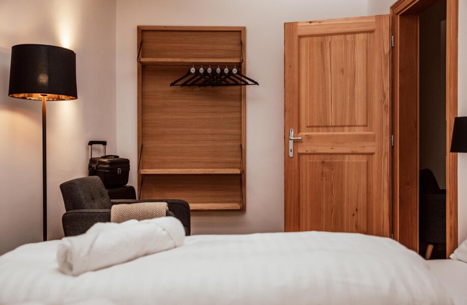 A cozy hotel room with a bed, wooden wardrobe, and open door. A floor lamp and suitcase are visible.