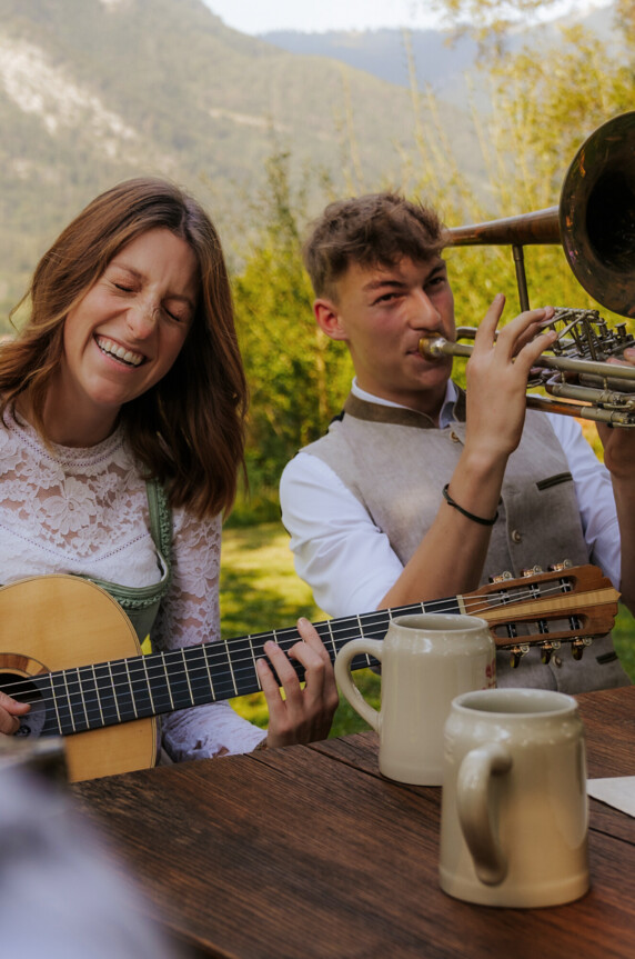 Zwei Personen musizieren fröhlich im Freien, eine Frau spielt Gitarre, ein Mann Posaune. Im Vordergrund stehen Bierkrüge auf einem Holztisch.