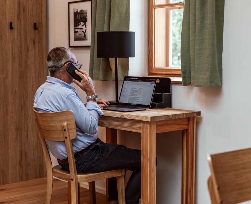 Ein Mann telefoniert in einem der Hotelzimmer, sitzt am Holzschreibtisch mit Laptop, neben Fenster mit grünen Vorhängen. Ideal für Workation in Salzburg.