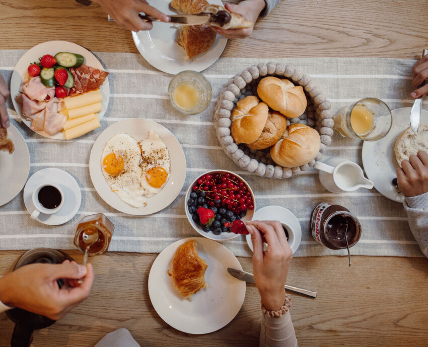 Reich gedeckter Frühstückstisch mit Spiegeleiern, Semmeln, Croissants, Aufschnitt, Käse, Beeren, Kaffee und Saft.