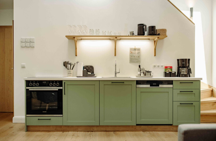 Modern kitchen with sage green cabinets, sleek appliances, and wooden shelves adorned with glassware.