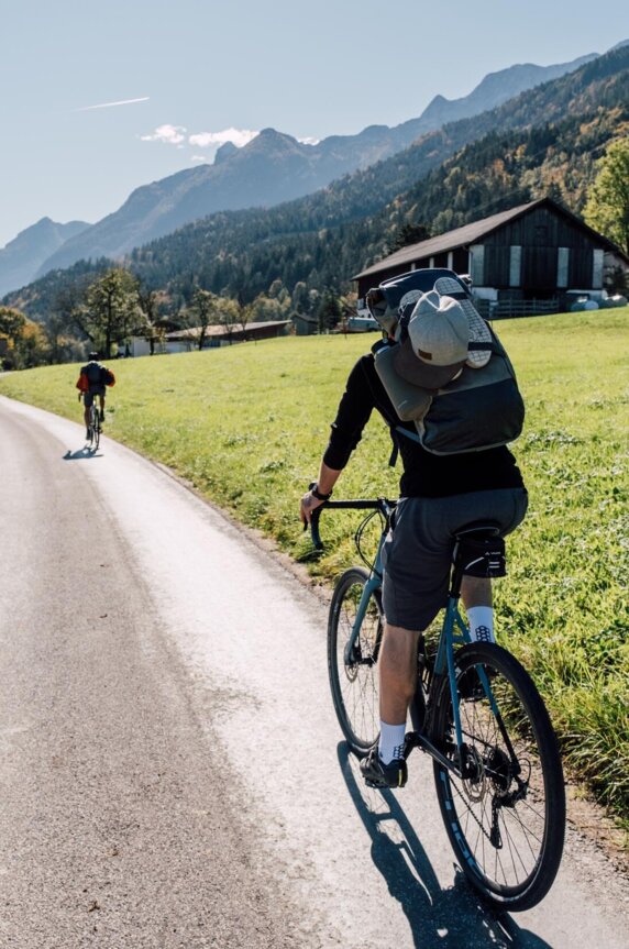 Zwei Radfahrer auf einem ländlichen Weg in einer malerischen Berglandschaft, mit grünen Wiesen und traditionellen Häusern.