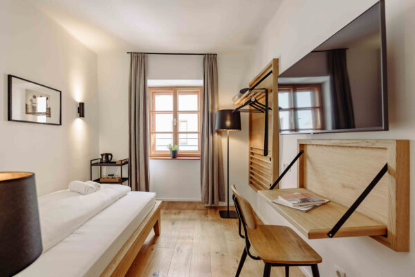 Modernes Hotelzimmer mit Einzelbett, Schreibtisch, Flachbildfernseher und großem Fenster mit Vorhängen, das eine gemütliche Atmosphäre bietet.