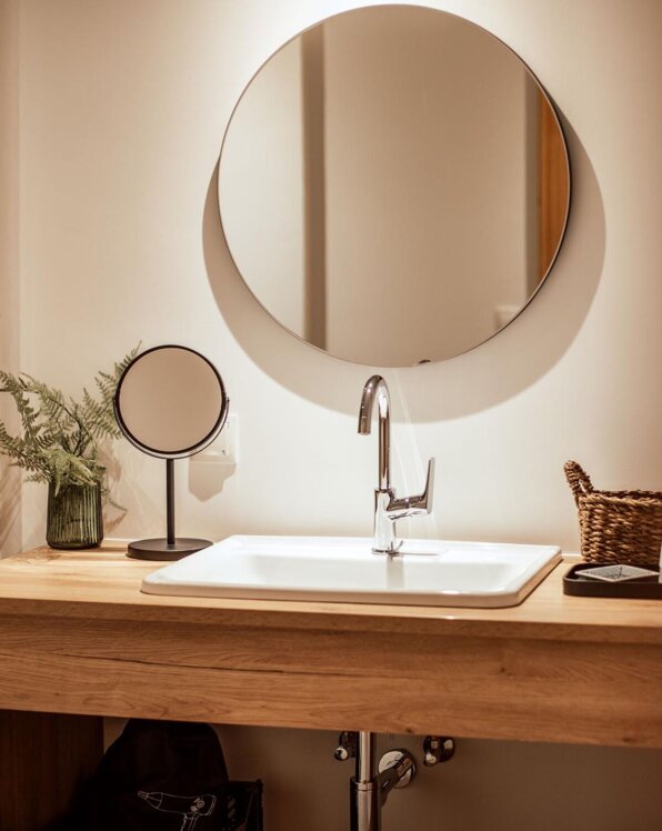 Modern bathroom with a round mirror, wooden countertop, sink, and decor including a plant and basket, creating a serene and stylish atmosphere.