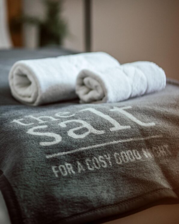 Weißes Handtuch, gerollt, liegt auf grauer Decke mit Aufschrift "the salt" in einem gemütlichen Zimmer.