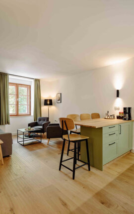 Modern living area with a cozy sitting space, wooden flooring, and a small kitchen with light green cabinets in a well-lit room.