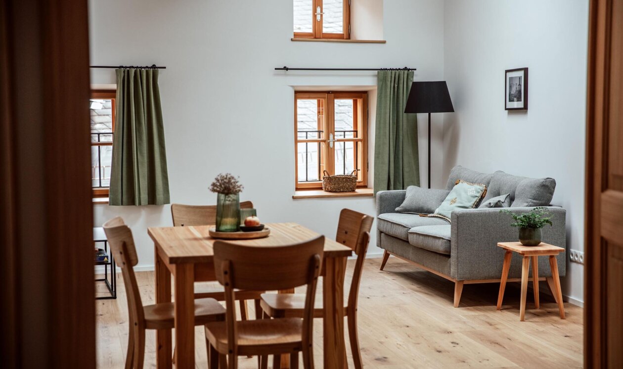 Gemütlicher Wohnbereich in einem Apartment von the salt Hallein in Salzburg mit Holztisch, grauem Sofa und grünen Vorhängen.