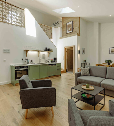 Modern living room with gray sofas, armchairs, a kitchenette with green cabinets, and decorative elements under a high ceiling with skylights.