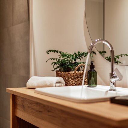 Moderner Badezimmer-Waschtisch mit grünem Pflanzenkorb, Handtuch und Seifenspender auf Holzunterlage vor einem großen Spiegel.