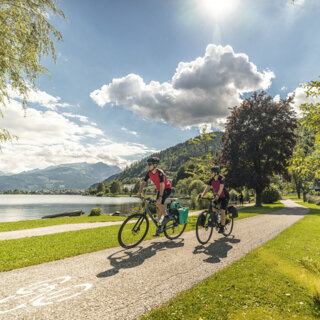 Zwei Radfahrer auf dem Tauernradweg am See, umgeben von grüner Landschaft und Bergen im Hintergrund.