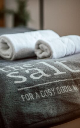 Aufgerollte Handtücher auf einer Decke mit dem Logo "the salt" in einem gemütlichen Zimmer.