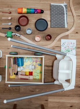 Spielzeuge und Kinderartikel auf Holzboden arrangiert, darunter eine Holzeisenbahn, bunte Becher, Löffel und ein weißer Hocker.
