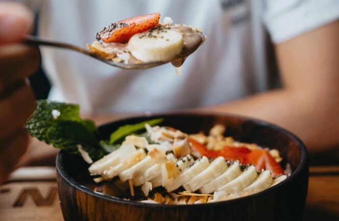 Smiling person holds a spoon over a wooden bowl of smoothie topped with banana, strawberry, chia seeds, coconut flakes, and mint.
